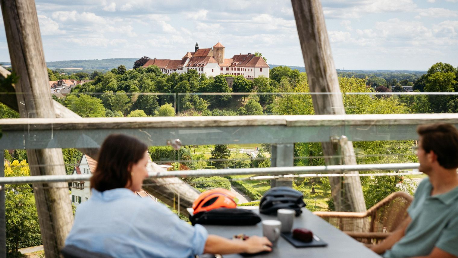 Blick von einem Café-Tisch über bewaldete Landschaft auf ein Schloss im Hintergrund.