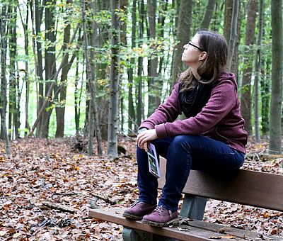 Auf diesem Foto sitzt eine braunhaarige junge Frau auf einer Parkbank und genießt mitten im Wald neben vielen Bäumen und bunten Herbstblättern den Gesundheitspfaden in Bad Essen. Dies ist ein Wanderweg, der zum Genuss mit allen Sinnen einlädt. Eine kleine Pause, um zwischendurch kräftig Durchzuatmen gehört auf einer Wanderung natürlich auch dazu. Nach der kurzen Pause geht es fit weiter mit dem Spaziergang. 