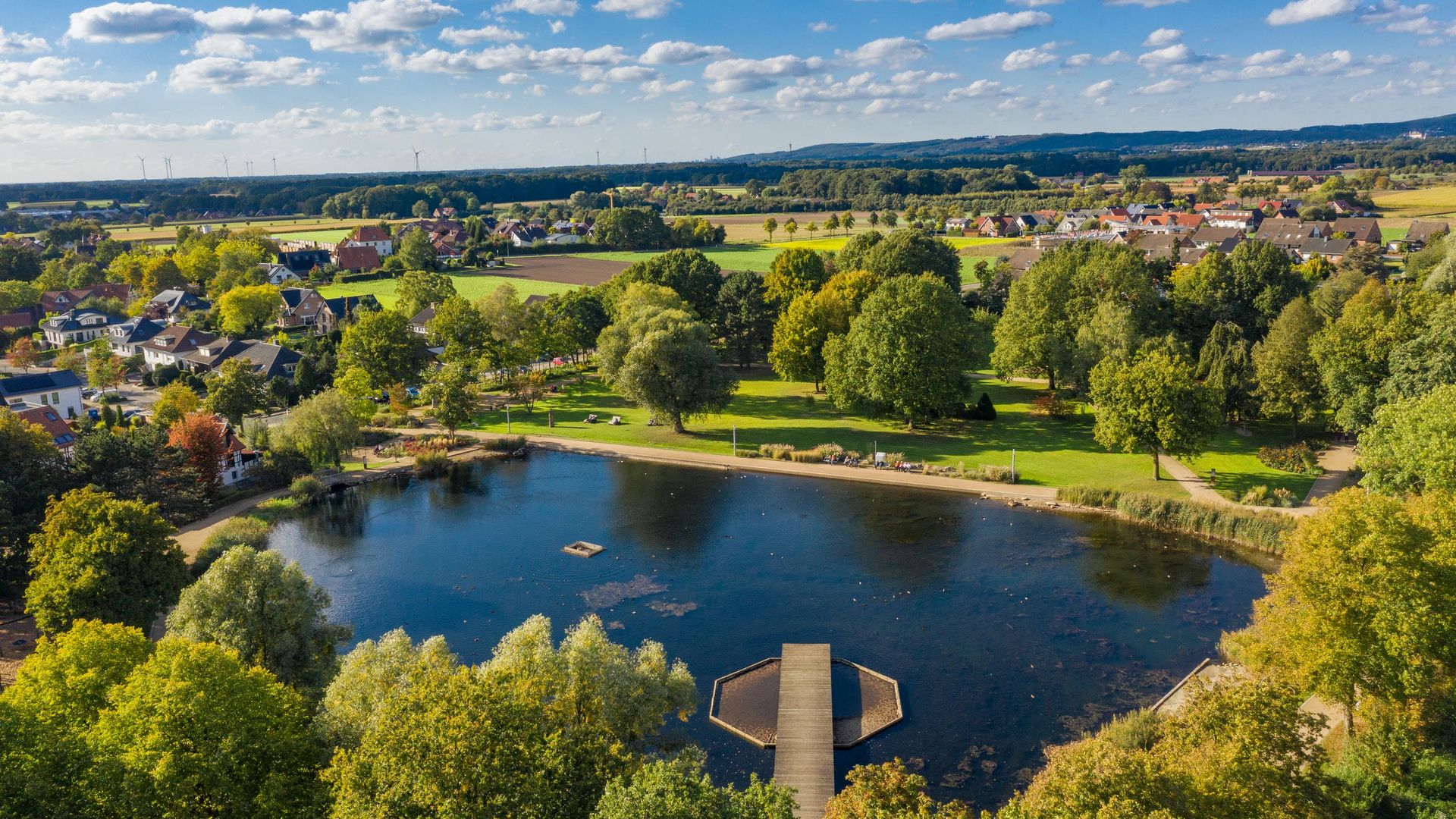 Landschaft mit See, Steg, grünen Bäumen und Dörfern im Hintergrund unter blauem Himmel.