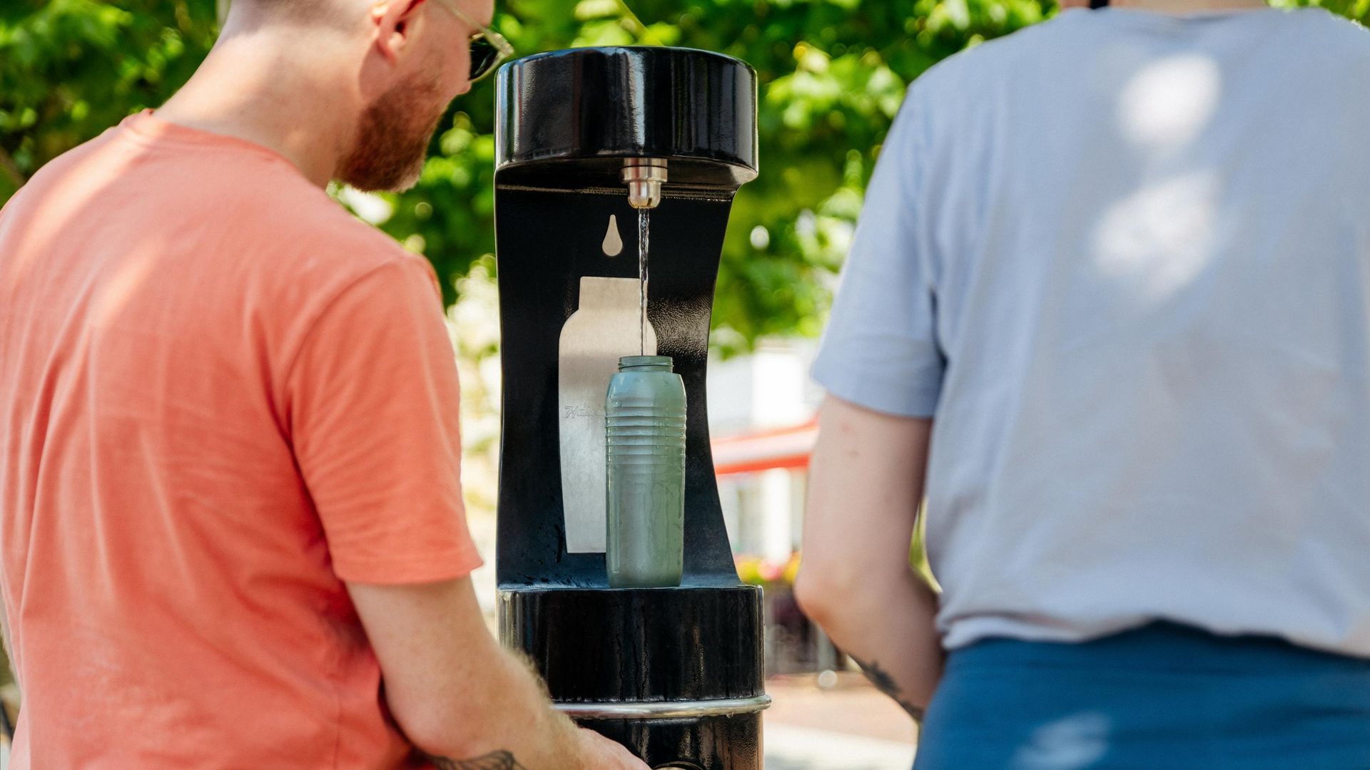 Zwei Personen füllen eine Flasche an einem öffentlichen Trinkwasserbrunnen nach.