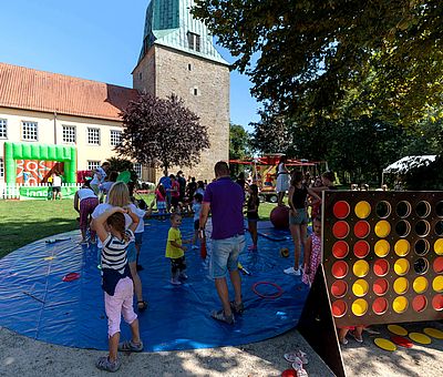 Beim Sommerfest der Kulturen in Fürstenau gibt es für Kinder viele Aktionen zum Mitmachen. Auf diesem Bild bekommt man einen Einblick von den verschiedenen Spielmöglichkeiten. Vorne rechts im Bild sieht man ein riesengroßes „Vier gewinnt“-Spiel stehen. Das Spielbrett besteht aus sieben Spalten (senkrecht) und sechs Reihen (waagerecht) in denen rote und gelbe Spielsteine stecken. Links im Foto, neben dem großen „Vier gewinnt”-Spiel ist eine große blaue runde Plane ausgelegt. Auf der Plane stehen Kinder und Erwachsene. Diese spielen mit unterschiedlichen Gegenständen, wie z.B. Keulen und Ringen. Hinter der blauen Plane sieht man eine grüne Rasenfläche. Weiter hinten auf dem grünen Rasen ist eine Hüpfburg aufgebaut. Diese ist mit einem weißen Zaun eingezäunt. Die Hüpfburg ist von außen grün und von innen rot. Hinter der Hüpfburg sieht man die Kirche stehen. Vor dem Kirchturm ist ein Kinderkarussell zu erahnen.