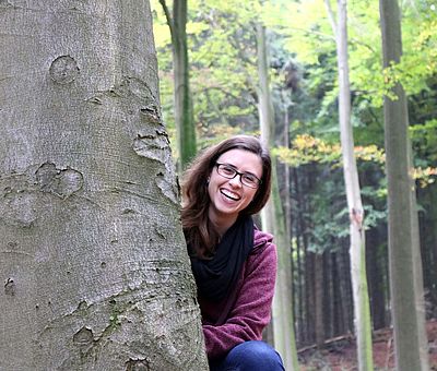 Auf einer Wanderung steht der Spaß im Vordergrund. Auf diesem Bild sitzt eine junge braunhaarige Frau hinter einem Baum und lacht in die Kamera. Im Hintergrund sieht man die herbstliche Natur mit bunten Blättern auf dem Boden und in den Bäumen. So macht die Wandertour über die Gesundheitspfade in Bad Essen allen Sinnen Spaß und hält fit. 