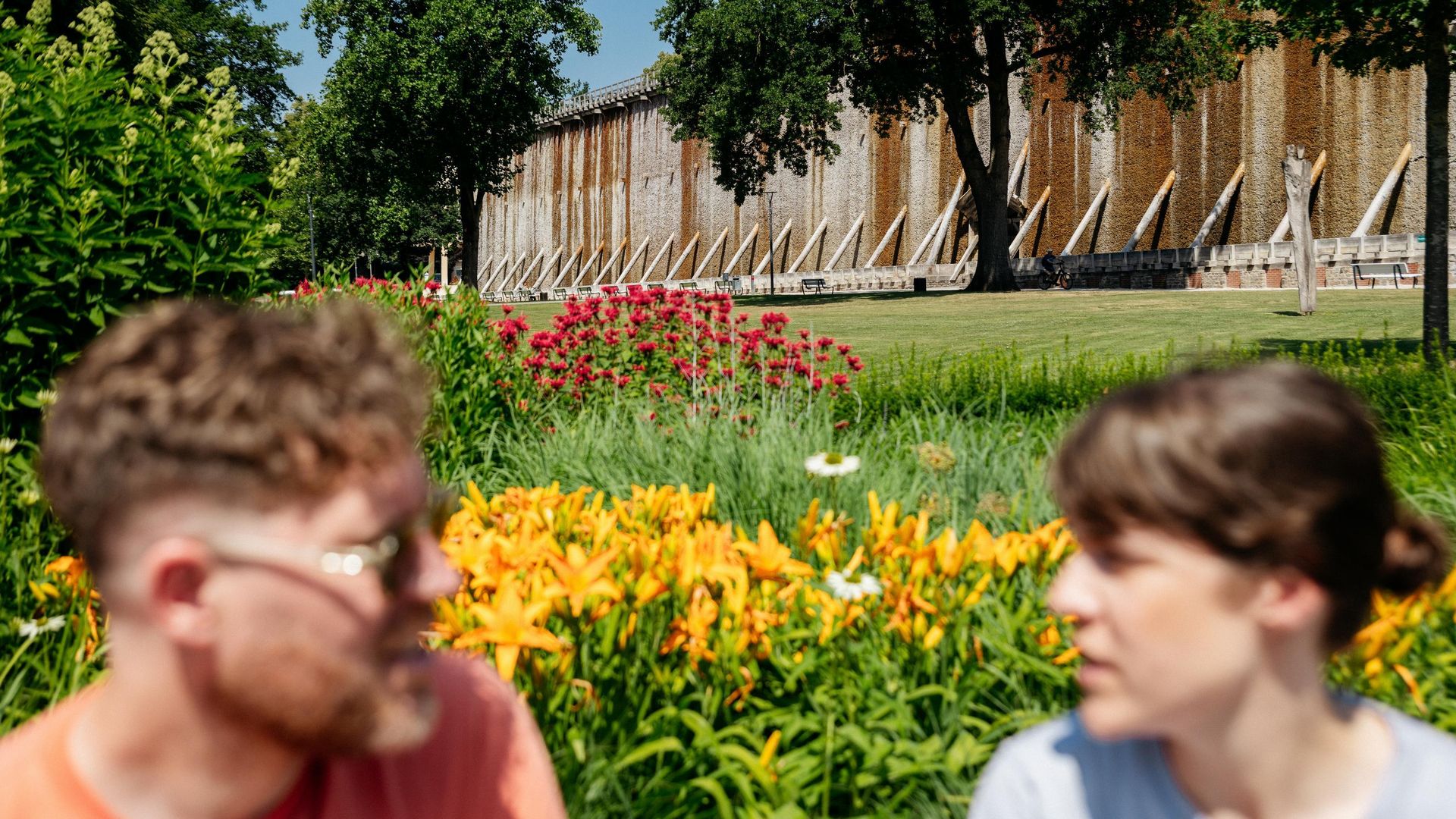 Zwei Personen vor einem alten Gradierwerk mit blühendem Garten im Vordergrund.