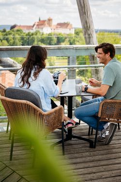 Zwei Personen genießen Kaffee auf einem Balkon mit Blick auf eine weit entfernte Burg.